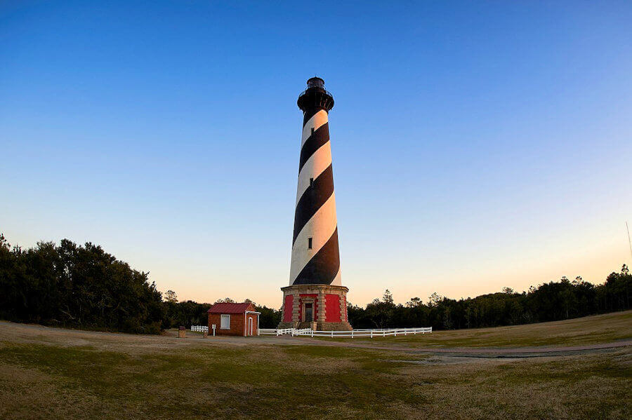 Black and white spiral striped light house with brick base sounded by a short white fence and a small building